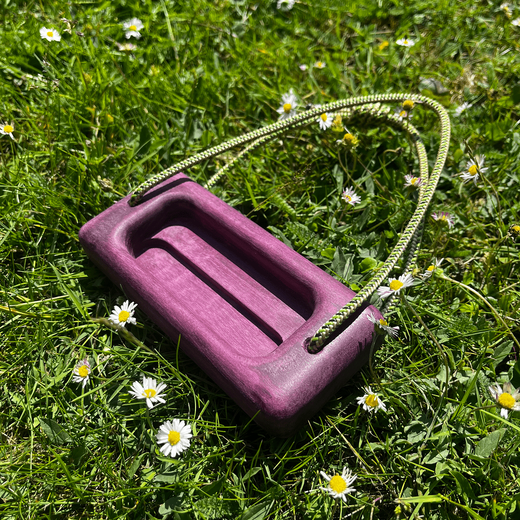 A micro hangboard made from purpleheart wood with green accessory cord, sitting in the grass with daisy's. the hangboard has rich purple hues.